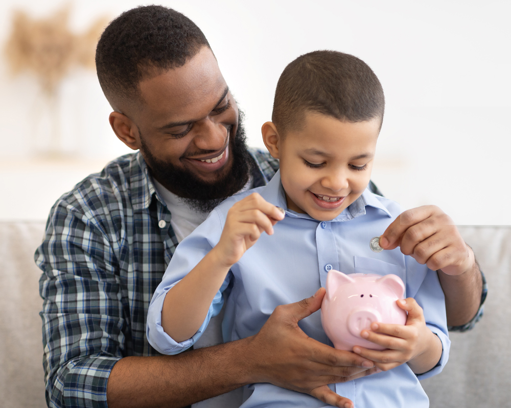 African Father And Son Putting Money In Piggybank At Home